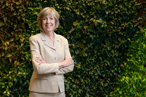 Dean Beth Walker stands with crossed arms in front of a wall of leaves