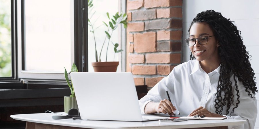 A woman writes notes as she studies something on her computer screen.