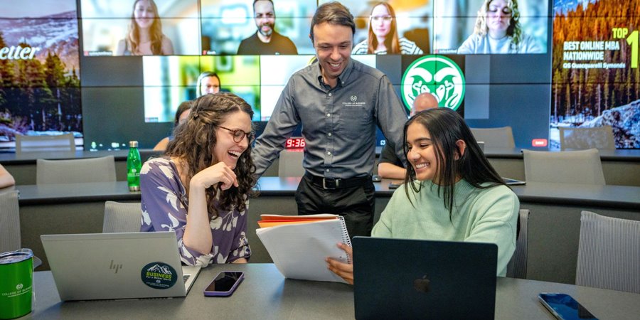 MBA students and a professor discuss an assignment in the Mosaic MBA classroom as virtually-participating students look on in the background.