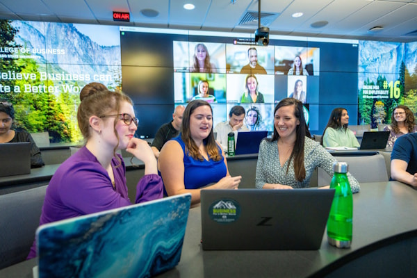 MBA students and a professor discuss an assignment in the Mosaic MBA classroom as virtually-participating students look on in the background.
