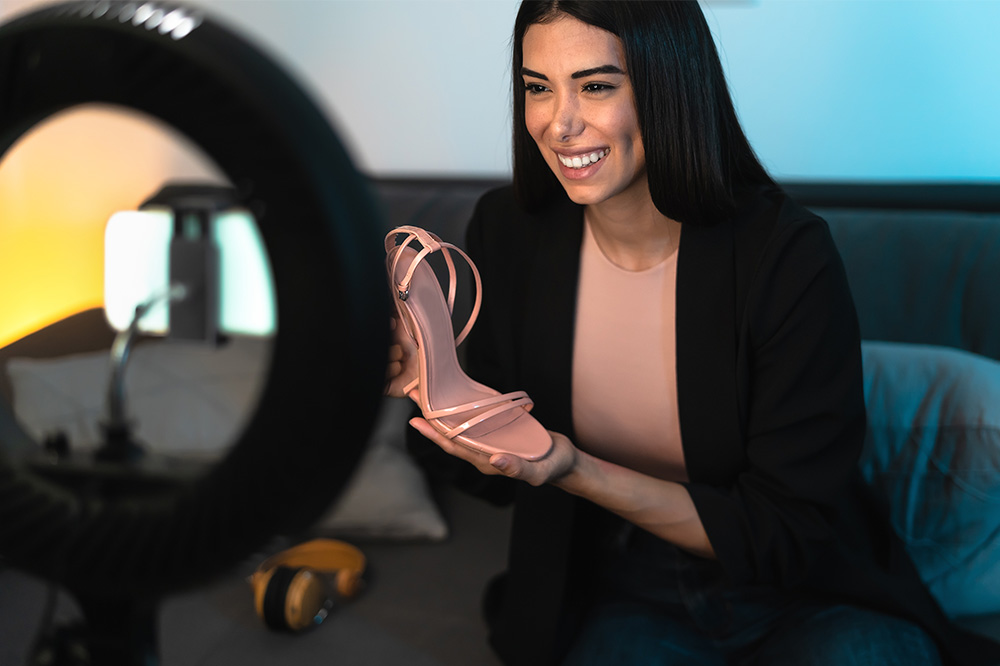 A woman holds a shoe and poses in front of a camera