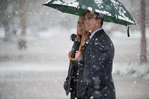 Two students walk through falling snow while holding an umbrella